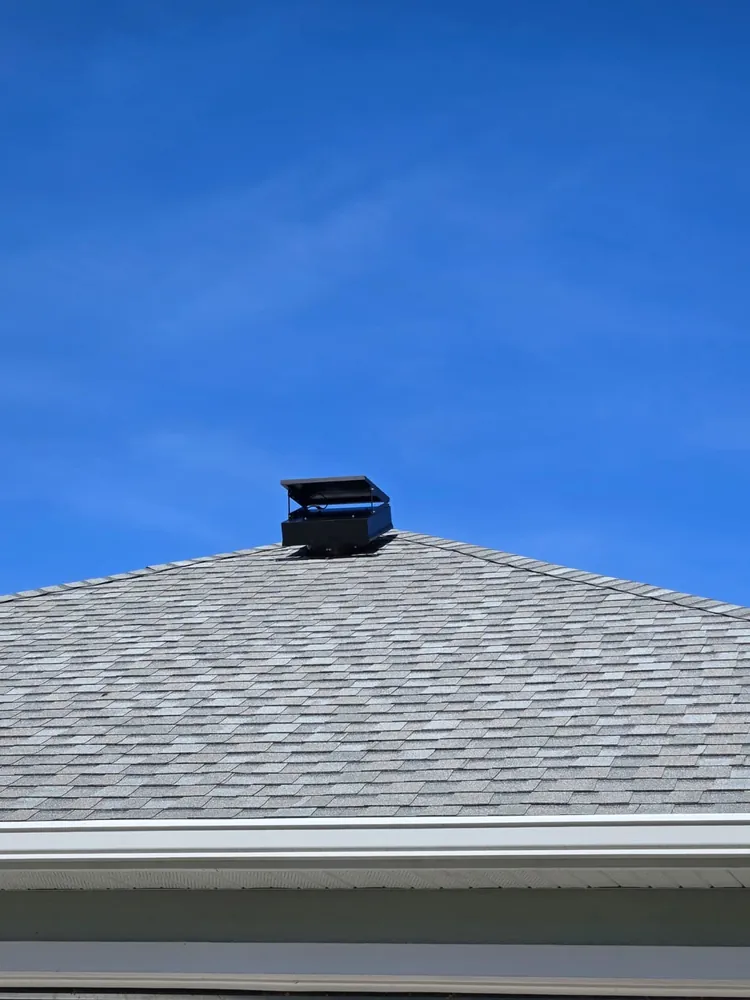 Low-profile solar attic fan at the peak of a grey shingle roof against blue sky
