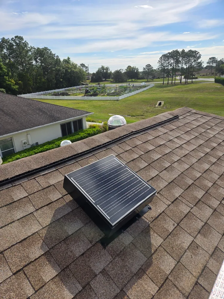 Low-profile solar attic fan on a brown shingle roof overlooking a community garden